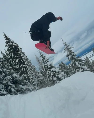 Alex airborne above snow-covered trees on a pink snowboard.