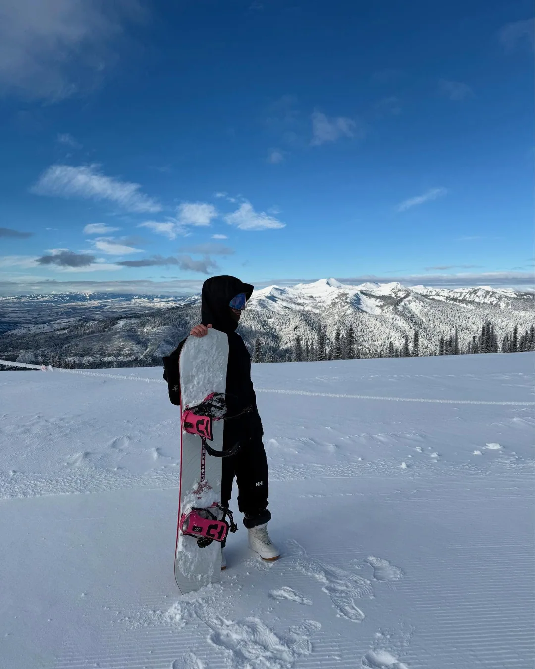 Alex holding his snowboard above a wide snowy mountain view.