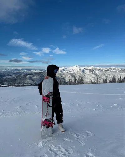 Alex holding his snowboard above a wide snowy mountain view.