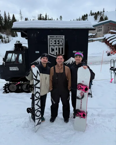 Three riders together with two snowboards beside a tracked vehicle.